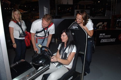 A group of four people engaged in a racing simulation experience at an event. One woman sits in a racing seat with a steering wheel, while two others guide her. A fourth person stands nearby, observing the scene.