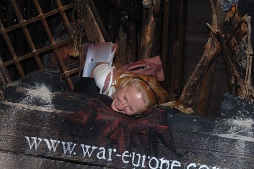 A woman with long blonde hair is playfully positioned on a wooden platform, surrounded by dark, rustic decor resembling a themed attraction. She is smiling while resting her head on a large sign displaying a web URL.