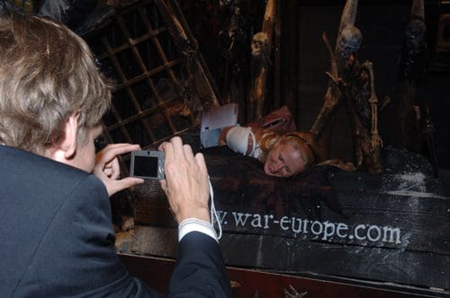 A man in formal attire taking a photograph of a woman who is lying on a dark, spooky-themed display with skeletal decorations. The background features ominous props, enhancing the eerie atmosphere.