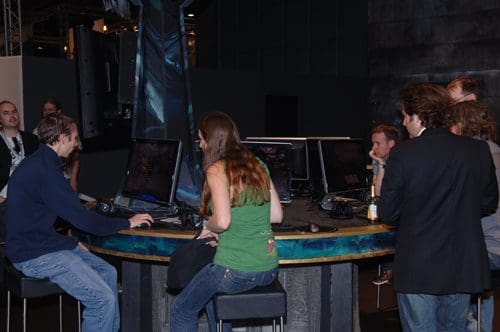 A group of individuals gathered around a modern gaming station with multiple screens. Two players are actively engaged at the center, while others observe or chat in a tech-inspired environment.
