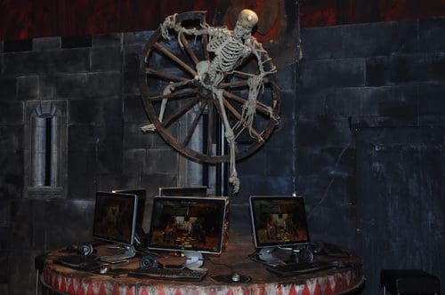 A skeleton suspended from a wooden wheel, positioned above several computer monitors with headsets, against a dark stone wall backdrop. The monitors display game graphics, contributing to a spooky atmosphere.