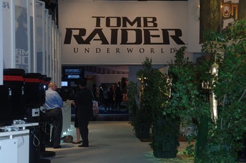 An exhibition space featuring a large sign that reads 'TOMB RAIDER UNDERWORLD' at the entrance. To the sides, gaming stations are set up with bushes adding greenery, and attendees are visible engaging with displays.