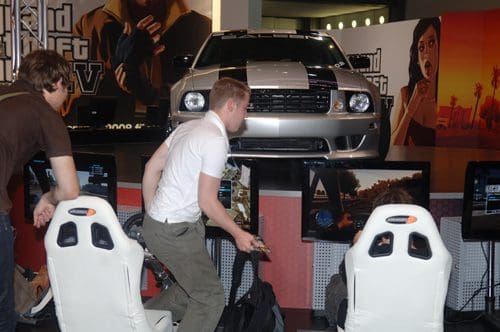 A gaming event featuring a large silver car on a stage. Two players are seated in white racing chairs, engaged in a video game, while a third person stands beside them, focused on the gameplay.