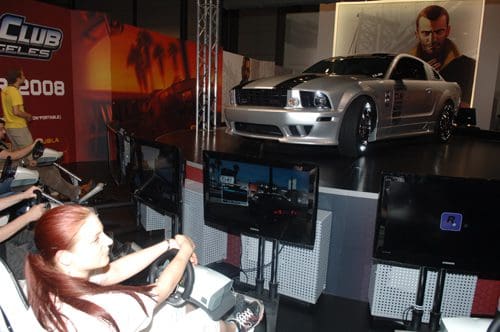 A silver sports car displayed prominently on a stage at a gaming event, surrounded by several gaming consoles and screens. A woman is seated in a racing simulator, focused on the screens, with vibrant graphics exhibited in the background.