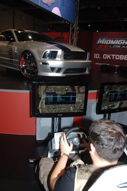 A person seated at a gaming setup, focused on screens displaying game information. In the background, a customized silver car with flashy wheels is elevated on a platform, adding to the vibrant atmosphere of the gaming event.