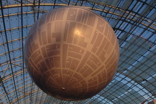 A large, spherical structure resembling a space station, suspended from a transparent ceiling. The design features geometric patterns and lights, creating a metallic appearance against the blue sky.