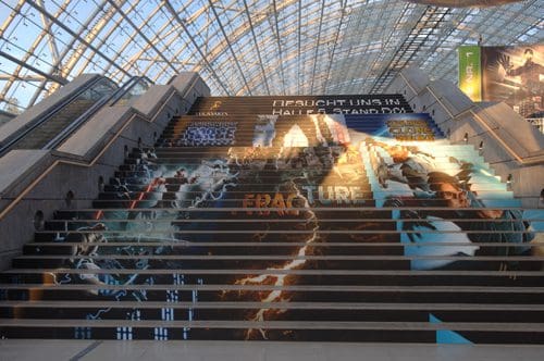 A large indoor staircase adorned with vibrant murals featuring various characters and designs, including lightning, faces, and the word "FRACTURE." Natural light streams in from a glass ceiling, enhancing the colorful artwork.