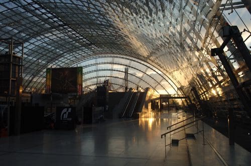 An expansive modern atrium with a large glass and metal roof reflecting sunlight. The interior features an empty space with escalators and posters, creating a bright and airy atmosphere.