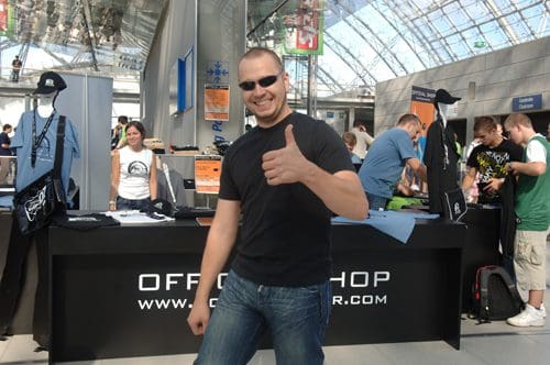 A man in a black T-shirt gives a thumbs-up while smiling in front of a merchandise table at a bright event. In the background, people browse through items and chat, with various shirts displayed.