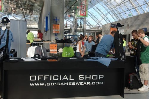 A display table at an event showcasing merchandise, including shirts and caps, with the label "OFFICIAL SHOP" prominently featured. Attendees browse items while engaging in conversation in a spacious, bright venue.