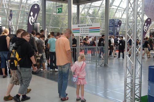A crowd of people, including a parent and a child, waiting in line at a registration point. The child wears a pink outfit and holds the parent's hand. The area is spacious with modern architecture and banners in the background.