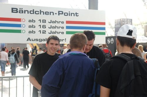 A group of young people chatting near a large sign labeled 'Bändchen-Point' with age requirements for entry displayed. The scene shows a bustling event atmosphere with attendees in the background.