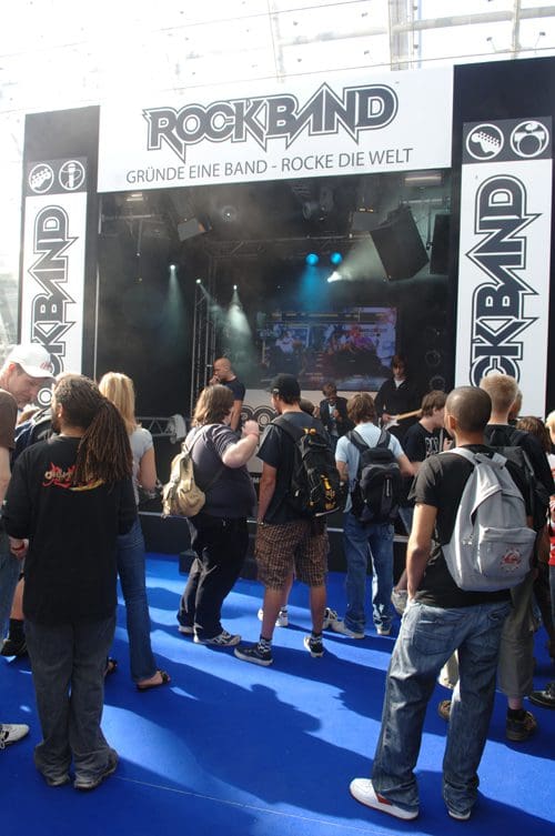 A crowd of people gathered in front of a stage at a Rock Band event, with a large banner displaying the Rock Band logo. The area is decorated with bright blue flooring and there are stage lights illuminating the scene.