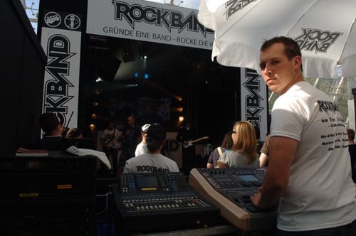 A sound technician monitoring audio levels at a live music event. Behind him, a band performs on stage, while attendees watch from a distance. The backdrop features a prominent "Rock Band" logo.