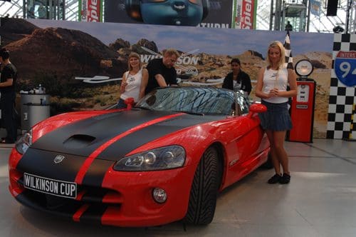A red sports car with black stripes prominently displayed in an exhibition space. Two women stand beside the car, wearing promotional outfits, while a man adjusts something under the hood. A large backdrop features a desert landscape.