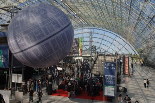 A large spherical balloon resembling a Death Star hangs above a bustling convention area filled with people. Stands and promotional banners are visible under a glass roof.