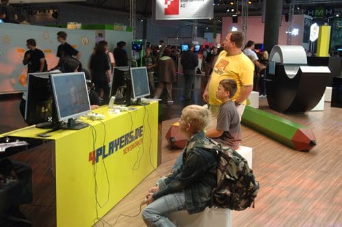 Two children seated in front of gaming monitors at an event, engaging in video games, while an adult observes nearby. The backdrop features vibrant booths and event attendees.