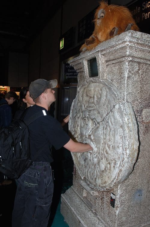 A visitor, wearing a gray hat and a black shirt, reaches towards a stone-like sculpture with an animal figure on top. The animal, resembling an orangutan, sits on a decorative pedestal amid a crowd in a large indoor space.