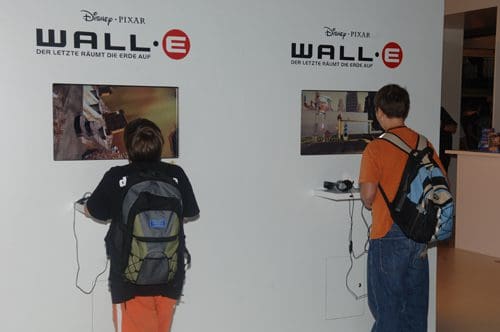 Two young boys engaged with interactive displays showcasing scenes from the animated film WALL-E. They wear backpacks and are focused on the screens mounted on a white wall, featuring vibrant visuals from the movie.