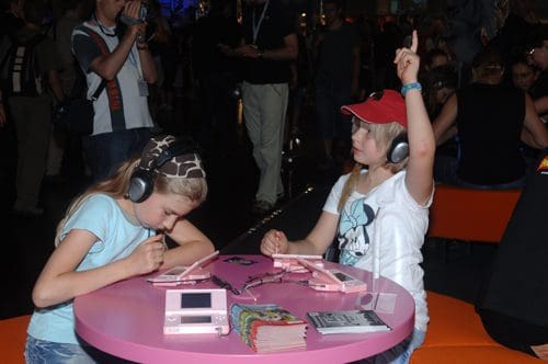 Two young girls sitting at a round pink table, each wearing headphones and playing handheld gaming consoles. One girl with a cap raises her hand excitedly while the other focuses on her game. People are in the background.
