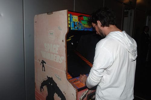 A person in a white hoodie playing a classic arcade game named 'Space Invaders.' The game cabinet features colorful graphics and a worn exterior, indicating its vintage style.