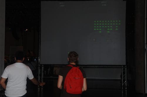 Two people standing in front of a large screen displaying a retro video game, with green graphics reminiscent of classic arcade games. One person wears a white shirt and the other wears a dark shirt with a red backpack.