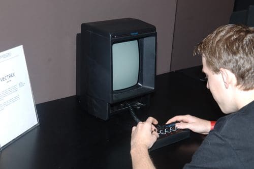 A person sitting at a table, playing on a black video game console with a joystick. The console has a small screen, and a sign is visible in the background, likely providing details about the device.