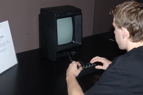 A person seated at a black table, focused on an old black-and-white computer monitor with a simple joystick controller. A sign describing the exhibit is visible on the left.