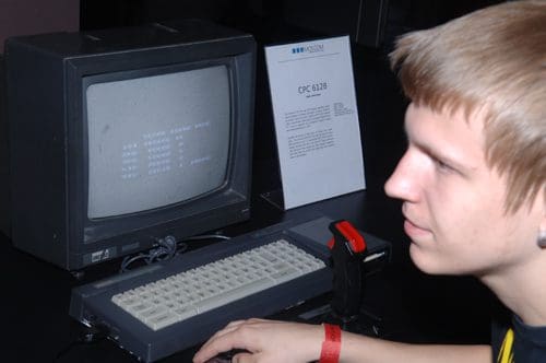 A young man focused on an old computer screen displaying text, with a retro keyboard and joystick beside him. A sign next to the computer reads "CPC 6128," providing information about the device.
