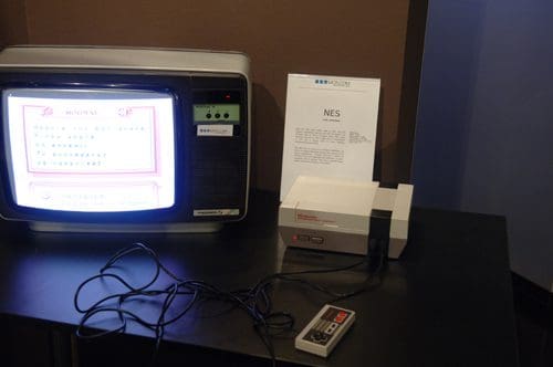 An old television displaying a classic video game on the screen sits next to a Nintendo Entertainment System (NES) console on a black table. A controller with buttons rests nearby, and an informational sign is positioned next to the console.