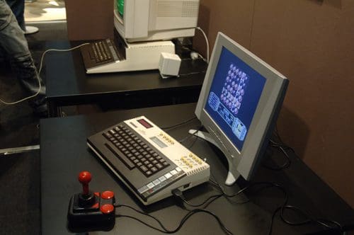 A retro computer setup on a black table, featuring a keyboard connected to a monitor displaying a colorful video game. An arcade joystick with red buttons is also visible, alongside another vintage computer in the background.
