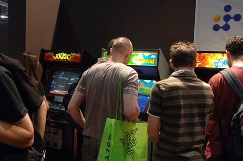 A group of people, mostly men, gathered around classic arcade game machines in a gaming event. Two arcade screens display colorful game graphics, while individuals engage with the games, showcasing a nostalgic atmosphere.