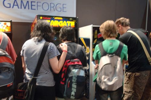 A group of four young people playing arcade games at a gaming event, standing in front of colorful arcade machines. The individuals have backpacks and are focused on the screens, creating an engaging atmosphere.