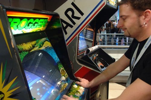 A person playing a classic arcade game at a gaming event, focused on the screen of the 'Frogger' machine, with a second arcade cabinet in the background displaying the Atari logo.