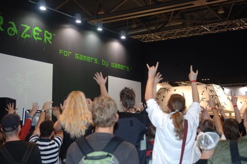 A crowd of enthusiastic event attendees raising their hands in excitement at a gaming expo, with a large display behind them featuring the Razer logo and tagline, "For Gamers, By Gamers."