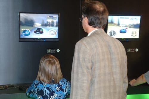 A man and a young girl stand in front of two screens displaying a racing video game. The girl, wearing a blue floral shirt, focuses intently on the game, while the man, in a grey suit, watches closely.