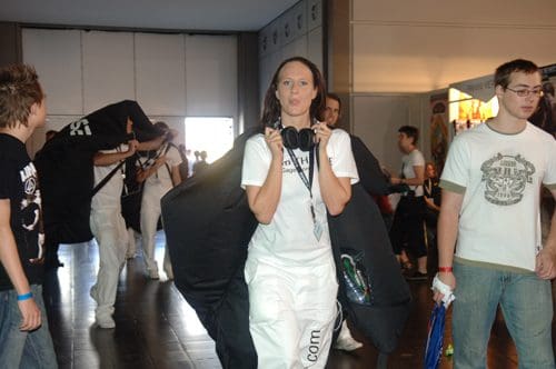 A group of individuals walking through a convention space, with one woman in the foreground holding headphones and wearing a white outfit. Others in the background are carrying large black bags.