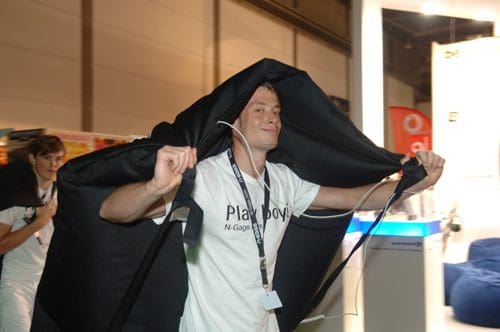 Two individuals are walking in a convention center, one wearing a large black cape-like garment, while the other appears to follow. The person in front has earphones and is wearing a white t-shirt with the word "Playboy" prominently displayed.