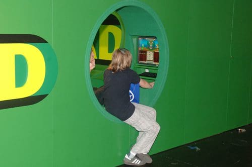 A child with long hair sits on the edge of a circular cutout in a green wall, reaching toward a gaming station visible inside. The setting is colorful, with hints of playful decor in the background.