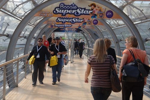 A busy walkway featuring attendees walking towards a sign that reads "Boogie SuperStar". People are carrying bags and wearing badges, moving through a modern glass corridor.