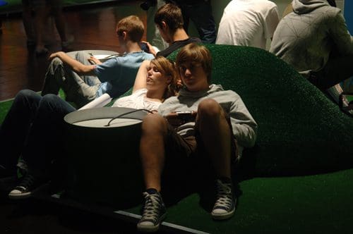 A young couple lounging together on a green, hill-shaped seating area, with the girl relaxing against the boy. In the background, other young people are seated, some looking at their devices.