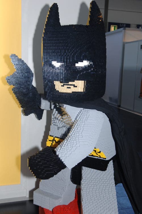 A life-size model of Batman made entirely from colorful LEGO bricks, positioned in an action stance with an extended hand. The figure features a black cowl, cape, and gray suit, set against a brightly lit background.