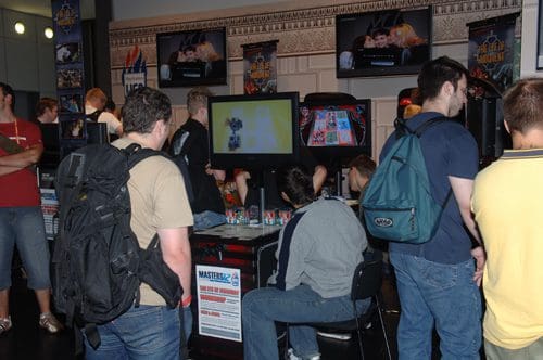 A group of young adults gathered around gaming stations at an event, with several screens displaying video games. Some players are focused on the games while others stand conversing, creating a lively atmosphere.