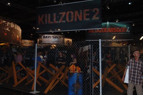 An exhibition booth for the video game "Killzone 2," featuring barricades made of wooden planks and a chain-link fence. Attendees are visible, exploring the setup and promotional displays in a busy convention atmosphere.