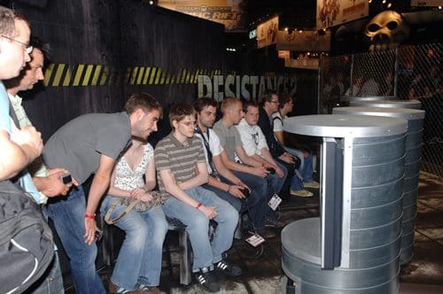 A group of gamers seated on benches, engaged in a multiplayer gaming experience at a convention. They are focused on screens in front of them, surrounded by a themed booth with industrial decor.