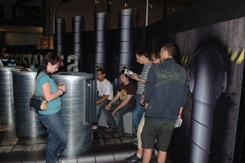 A group of five people interacting at a promotional event, with two seated at a booth while three others, including a woman in a striped shirt, engage with a display featuring themed decorations.