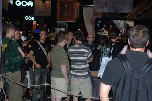 A crowd of people gathered in front of large screens displaying video games at a convention. Attendees include young adults dressed casually, engaged in conversation while looking at the screens. The environment is vibrant, filled with gaming-themed decorations.