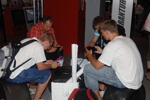 Four young men sitting on large black tires, focused on handheld gaming devices. A man in a plaid shirt watches them while seated nearby. The backdrop features a red wall with a partially visible sign.