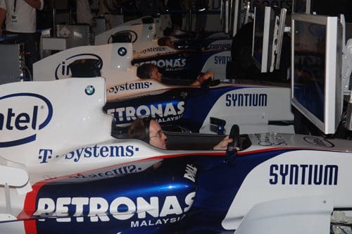 Two racing simulator setups featuring Formula 1 car replicas, where participants are seated with steering wheels, immersed in a driving experience. The cars are adorned with sponsor logos, including Petronas and Intel.