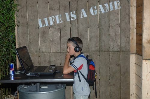 A young boy wearing headphones stands in front of a laptop at a table, concentrating on a screen. Behind him is a wooden wall with the phrase "LIFE IS A GAME" painted in white. He has a small backpack on his shoulders.